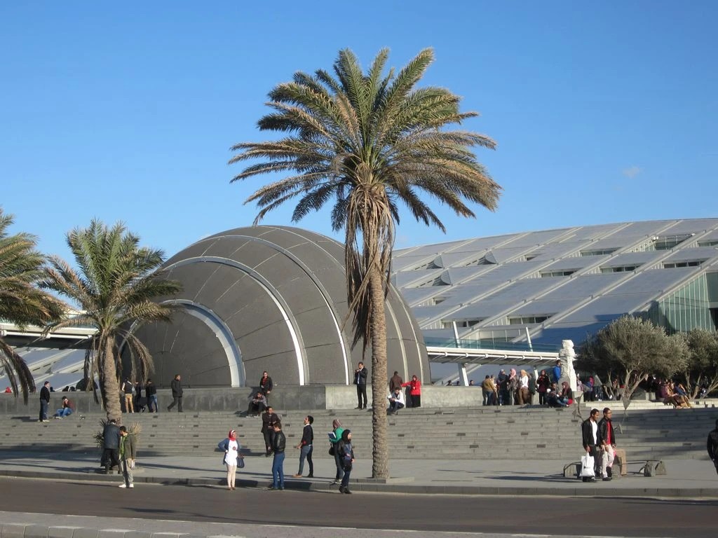 The Bibliotheca Alexandrina planetarium dome and sloping glass roof, with palm trees and visitors on the front steps